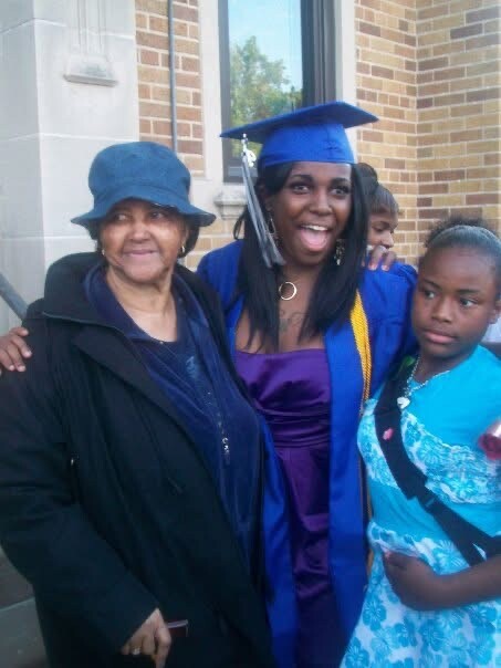 Graduate in blue cap and gown posing with an older woman and a young girl outside a brick building, all smiling.