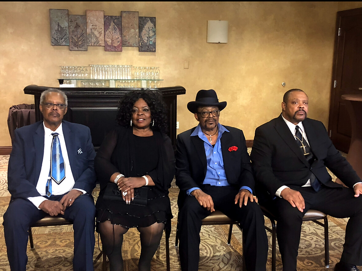 Four adults in formal attire seated in a row in a beige hotel lounge with a piano behind them.