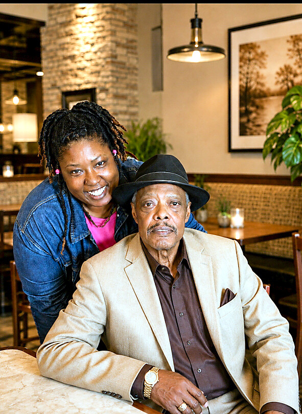 Smiling woman stands behind an older man in a beige blazer at a restaurant table.