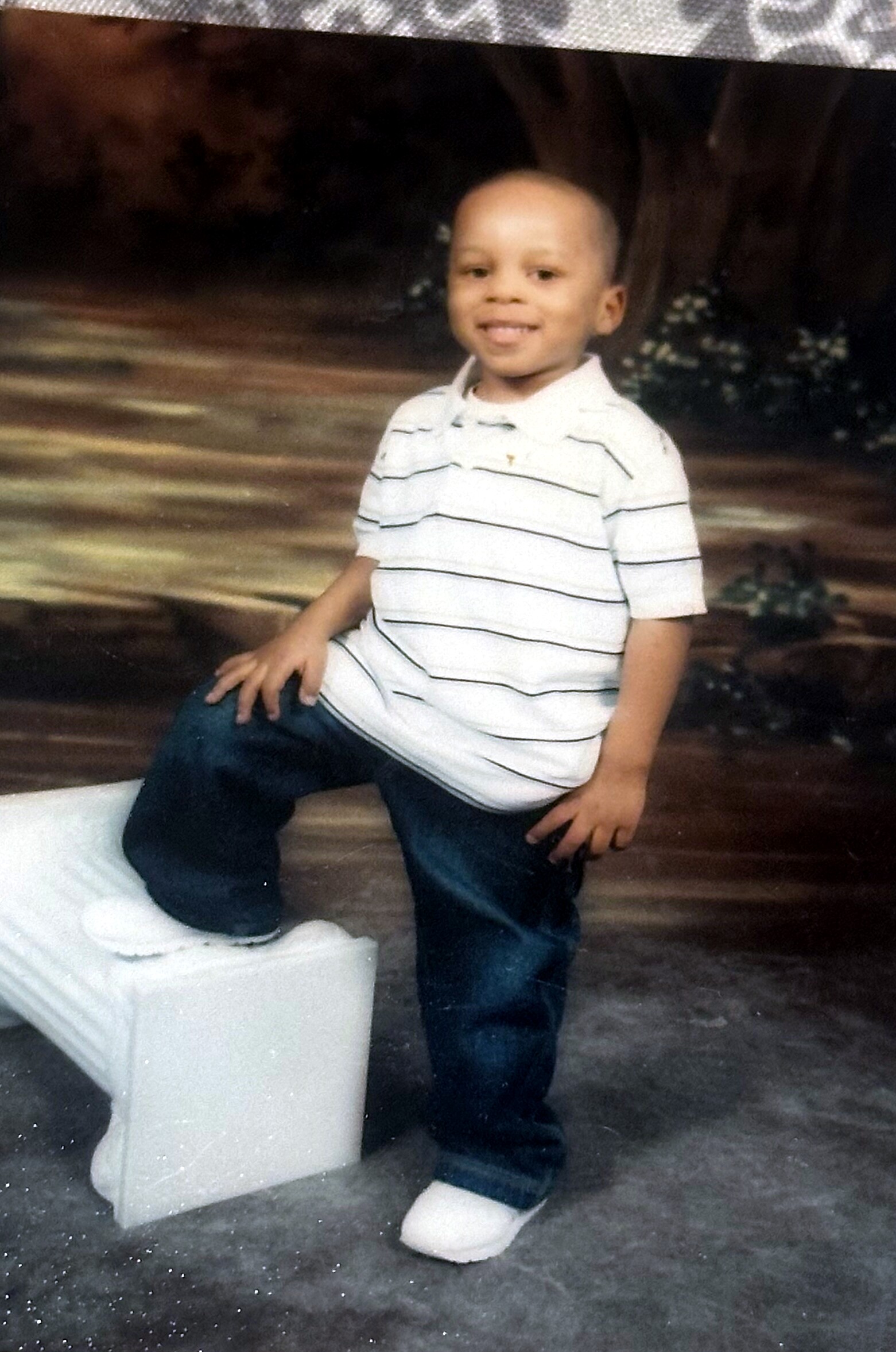 Smiling young boy in a striped polo shirt and jeans posing beside a white prop in a studio-like outdoor backdrop