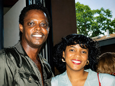 A man and a woman smiling at the camera, standing together near a doorway with greenery visible outside.