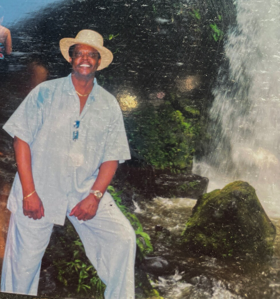 Smiling man in a light blue shirt and pants wearing a straw hat stands beside a rocky waterfall in a tropical setting.