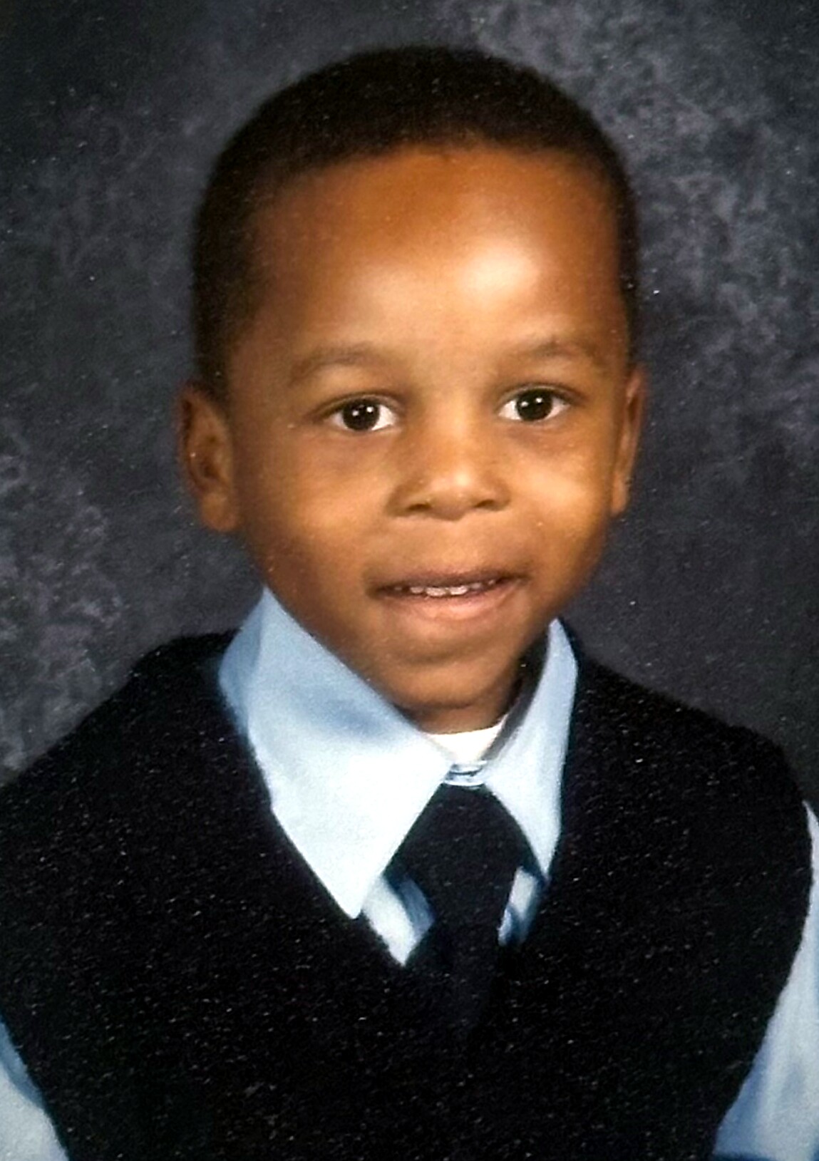 Young boy in a light blue shirt and dark sweater vest posing for a school portrait, smiling slightly.