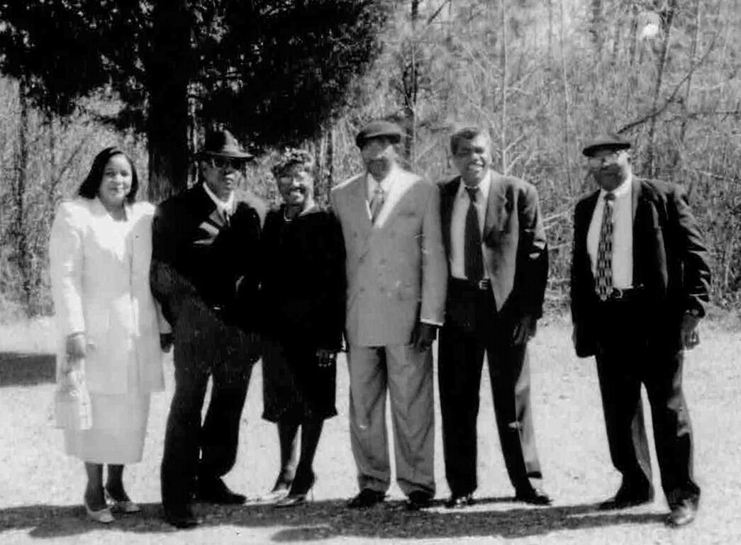 Seven adults in formal attire posing outdoors in a sunny, wooded area.