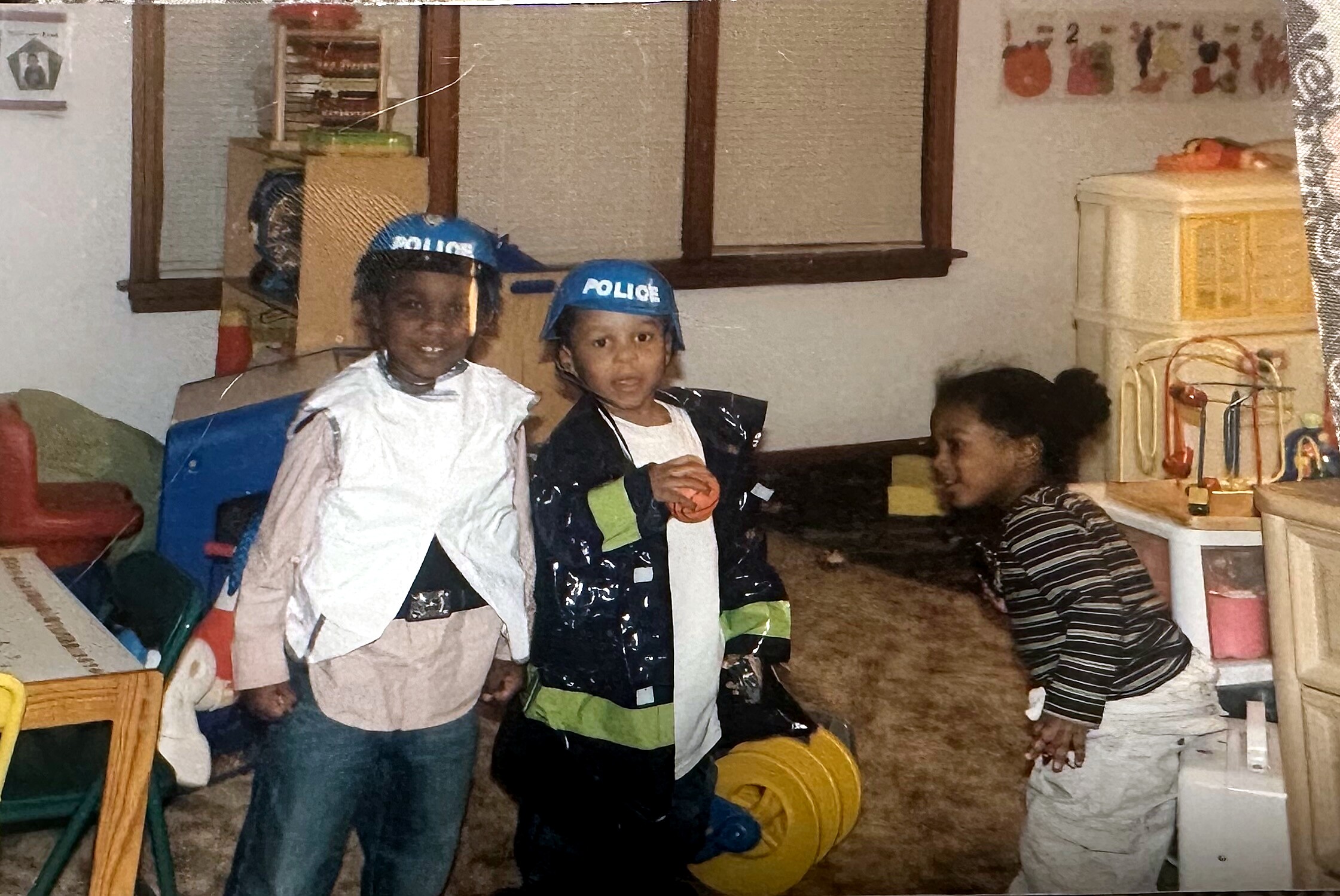 Two children in homemade costumes posing in a cluttered playroom, one wearing a blue police helmet, with a girl in a striped shirt seated nearby and assorted toys around.