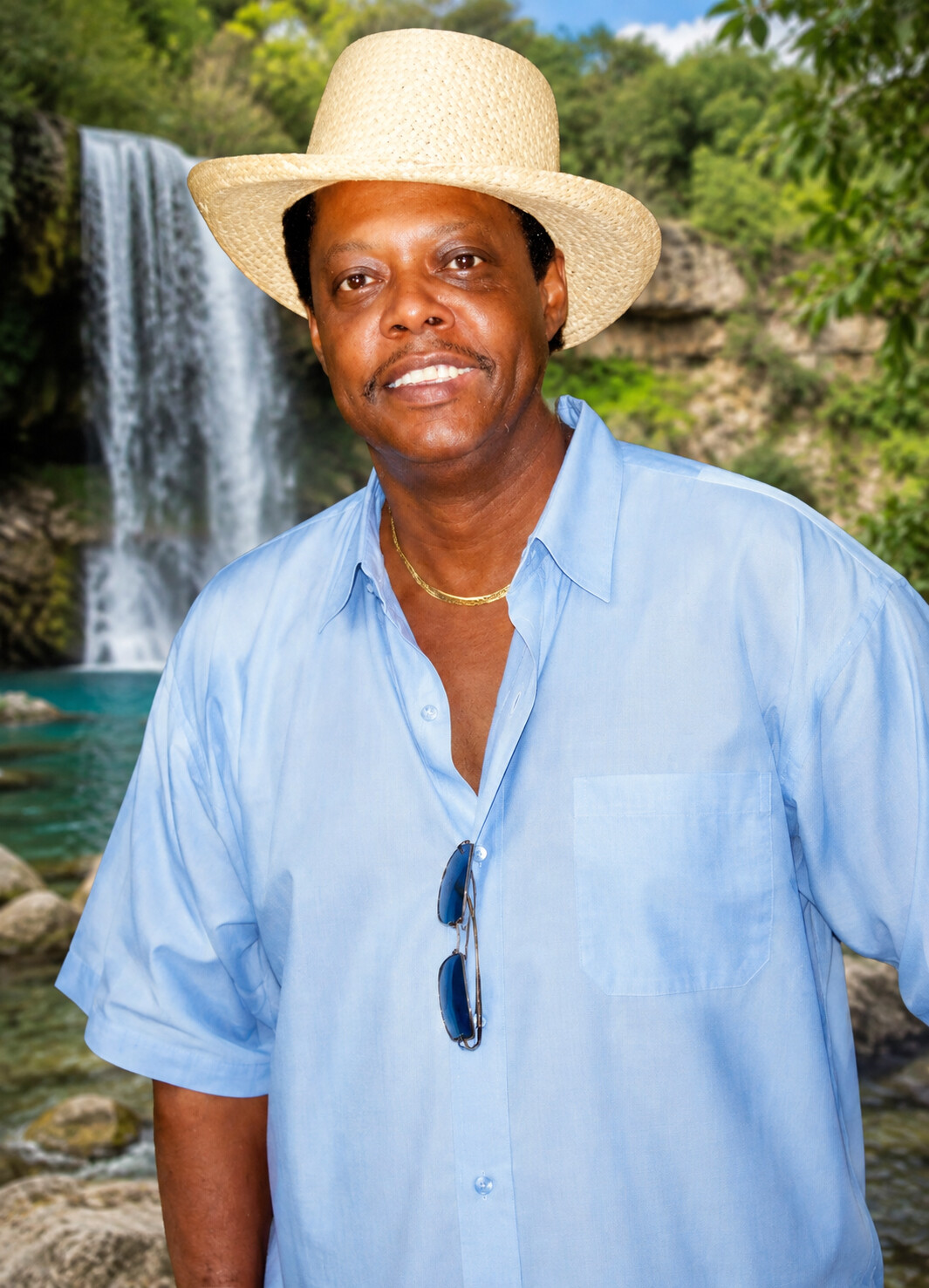 Smiling man in a straw hat and light blue shirt posed outdoors near a waterfall and greenery, sunglasses hanging from his shirt pocket.
