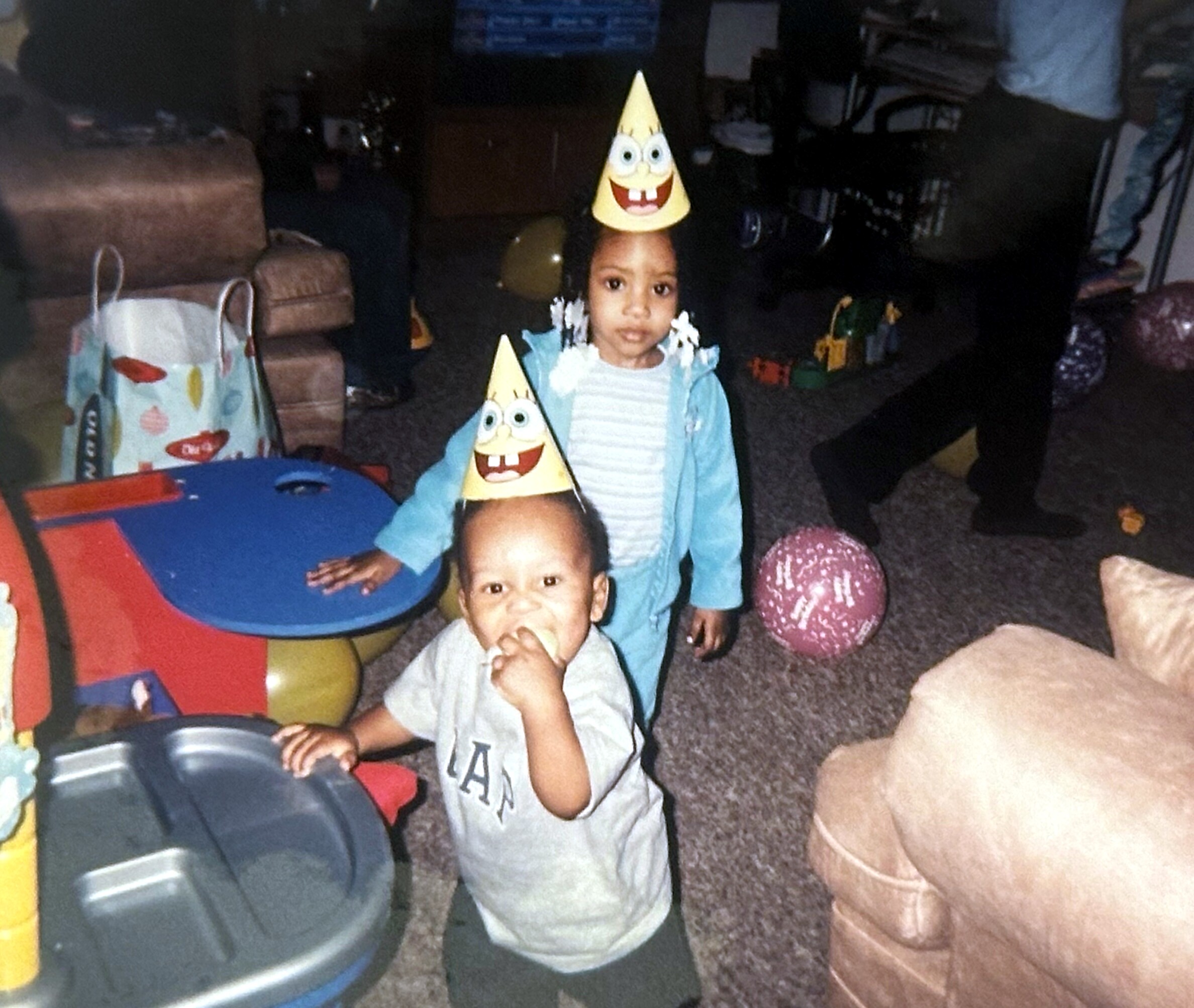 Child wearing a cone-shaped party hat with a cartoon face at a birthday party, with toys and a play area in the background.