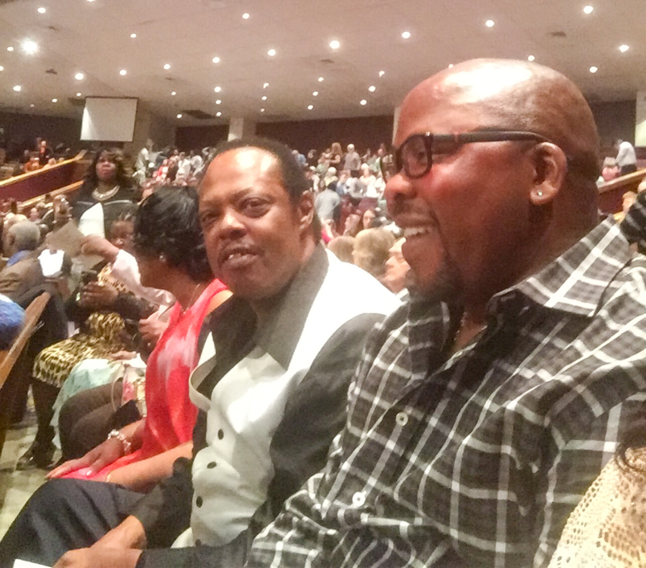 Two men seated in a crowded auditorium, smiling, with other attendees visible in the background.