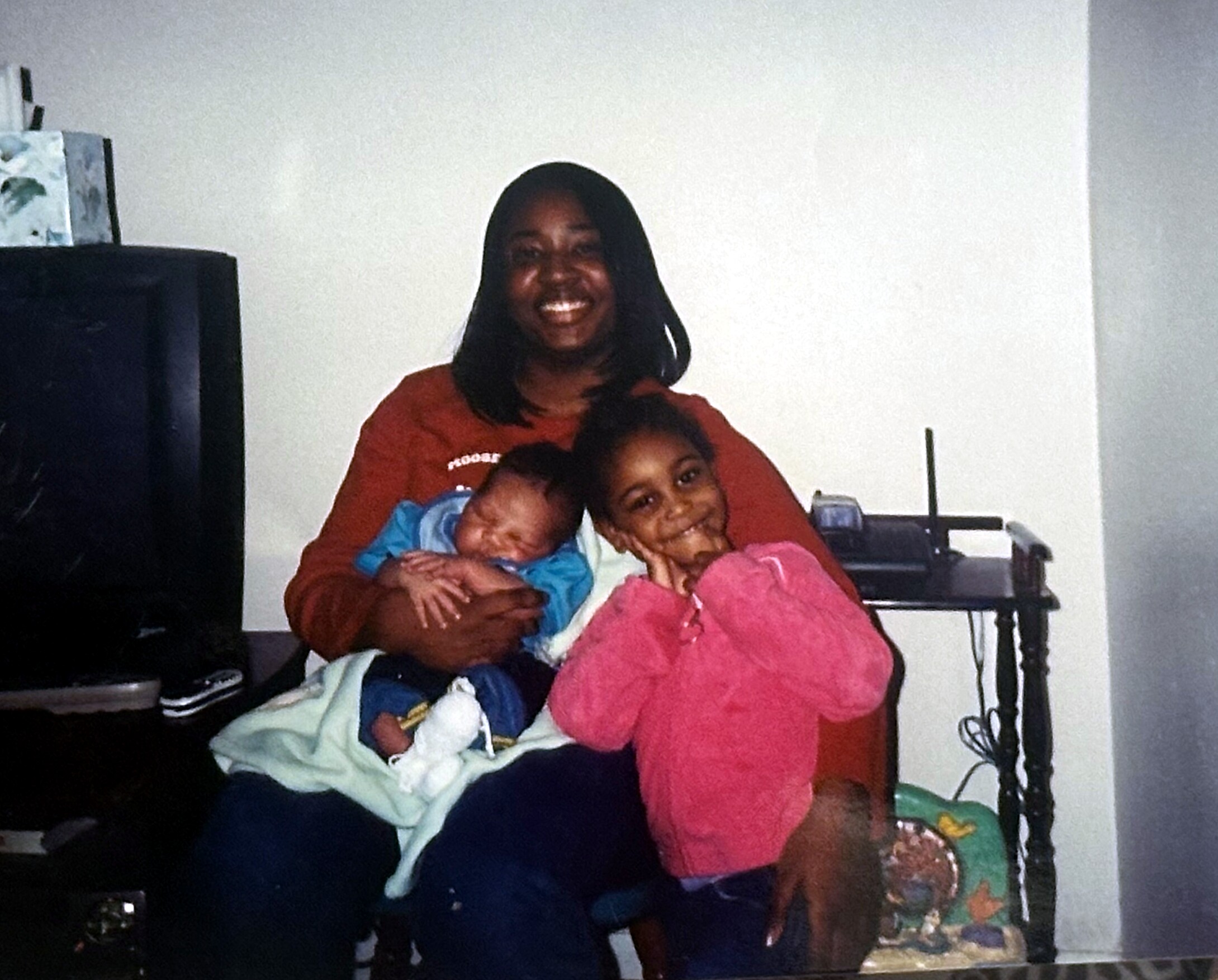 Smiling woman holding two young children wrapped in blankets indoors in a cozy home setting.
