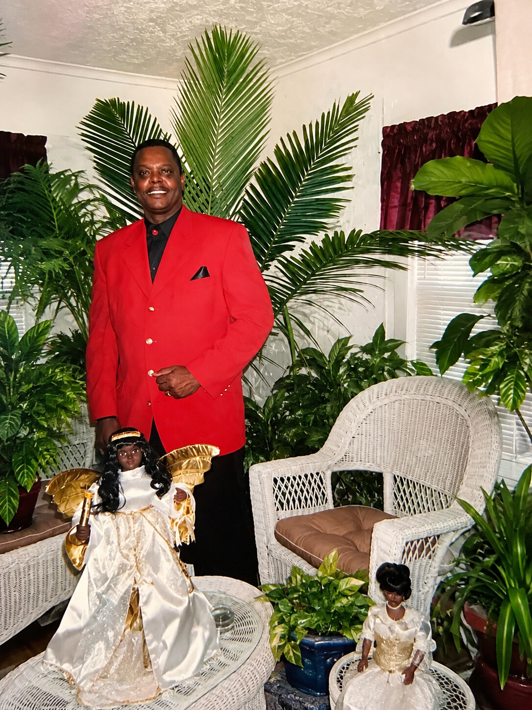 Man in a red blazer stands smiling beside two ornate dolls among lush indoor plants.