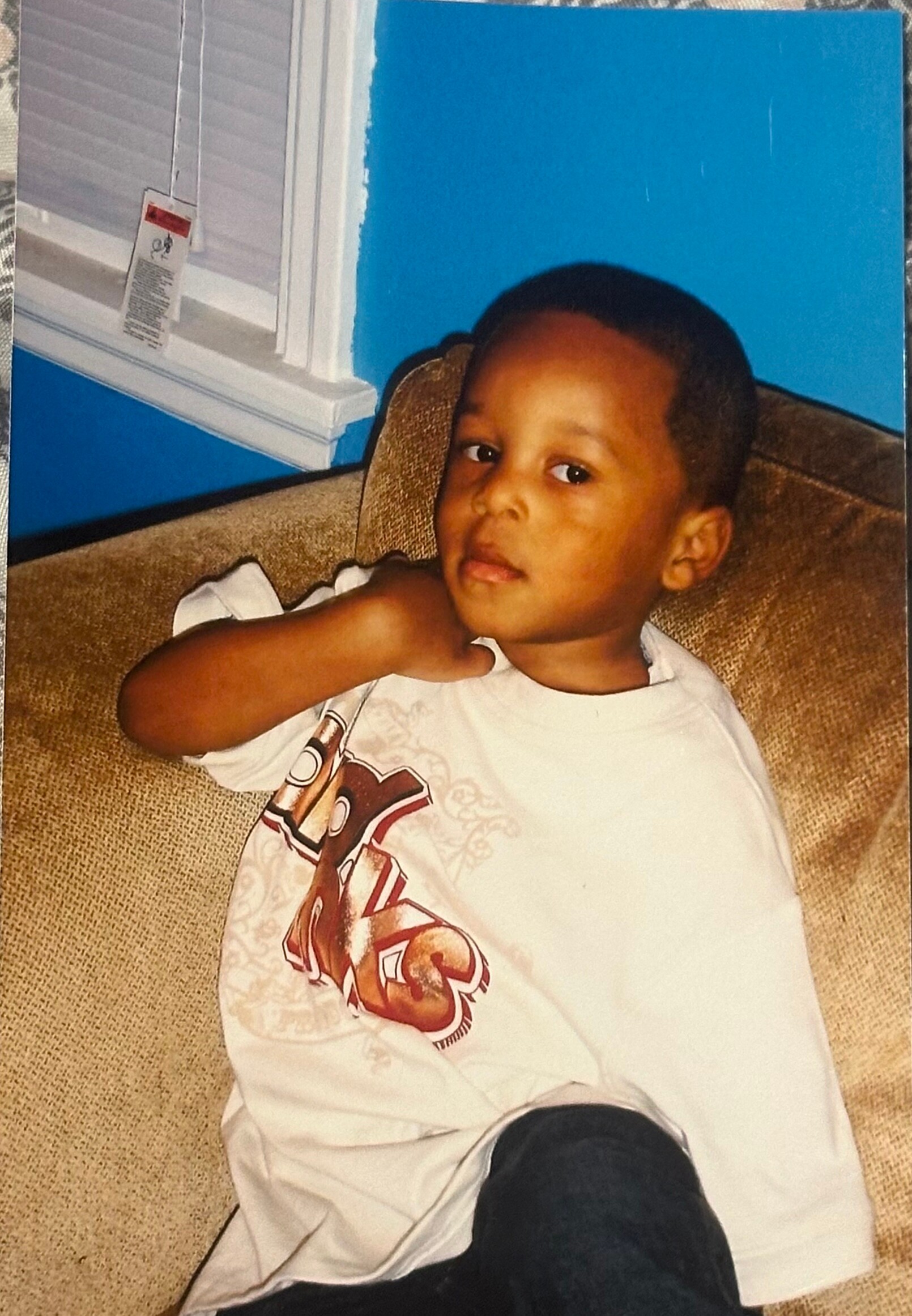 Young child sitting on a brown couch, wearing a white printed T-shirt, with a blue wall and window blinds in the background.
