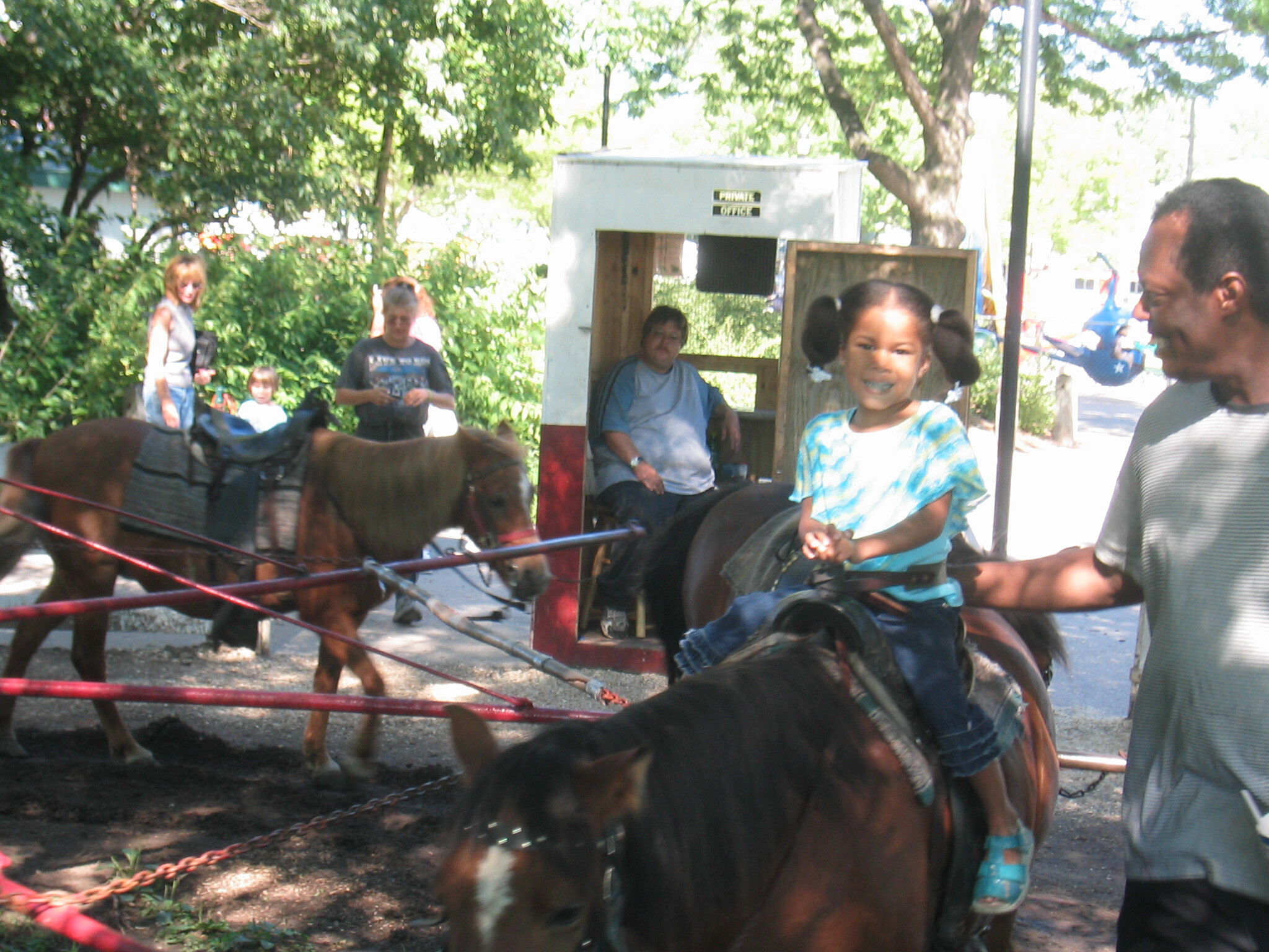 Young girl smiles while riding a pony, guided by a man nearby, with a white booth and trees in the background.