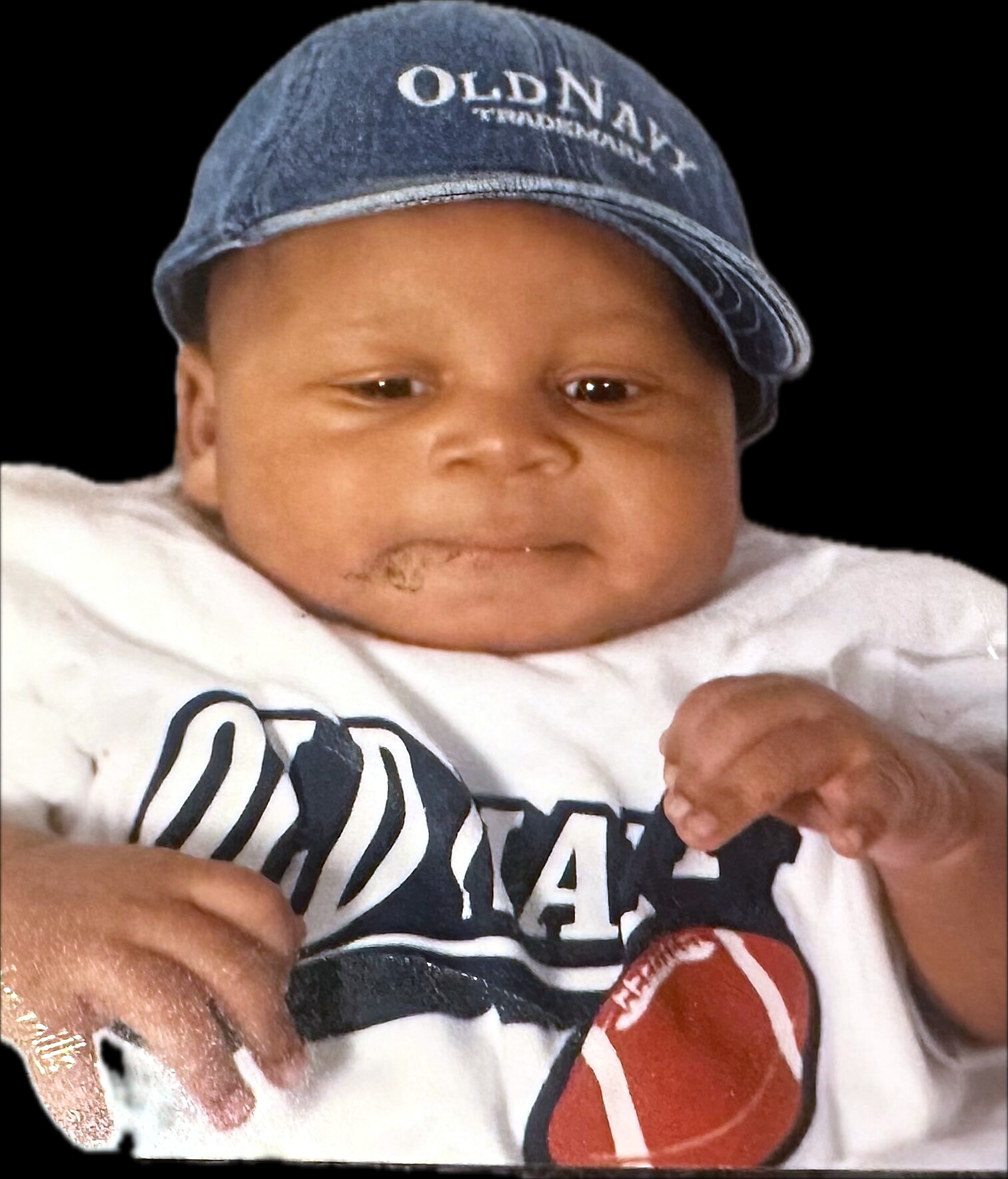 Close-up of a baby wearing a faded blue cap and white shirt, against a black background, looking toward the camera.