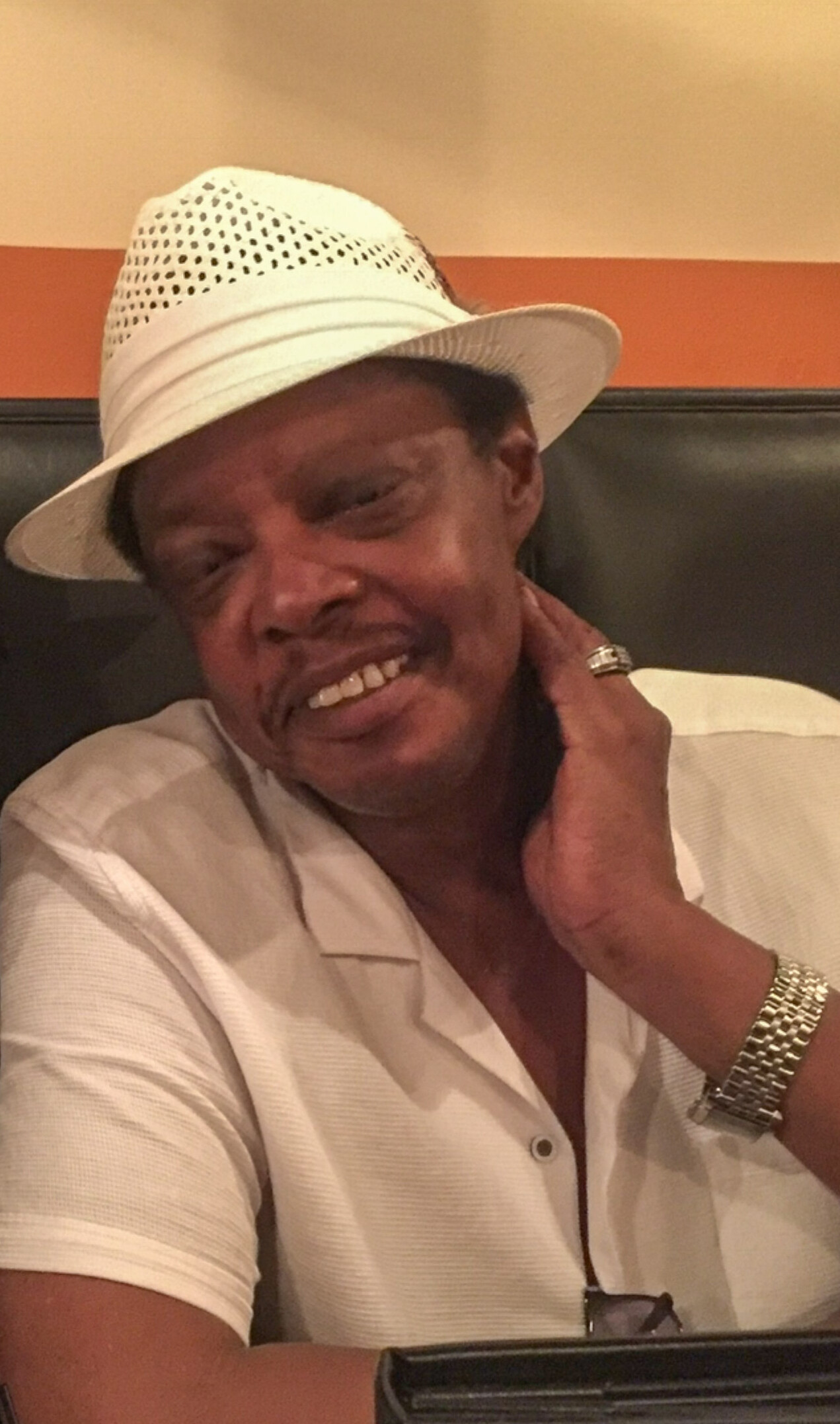 Smiling man in a white fedora hat and light shirt, resting his head on his hand in a restaurant booth.