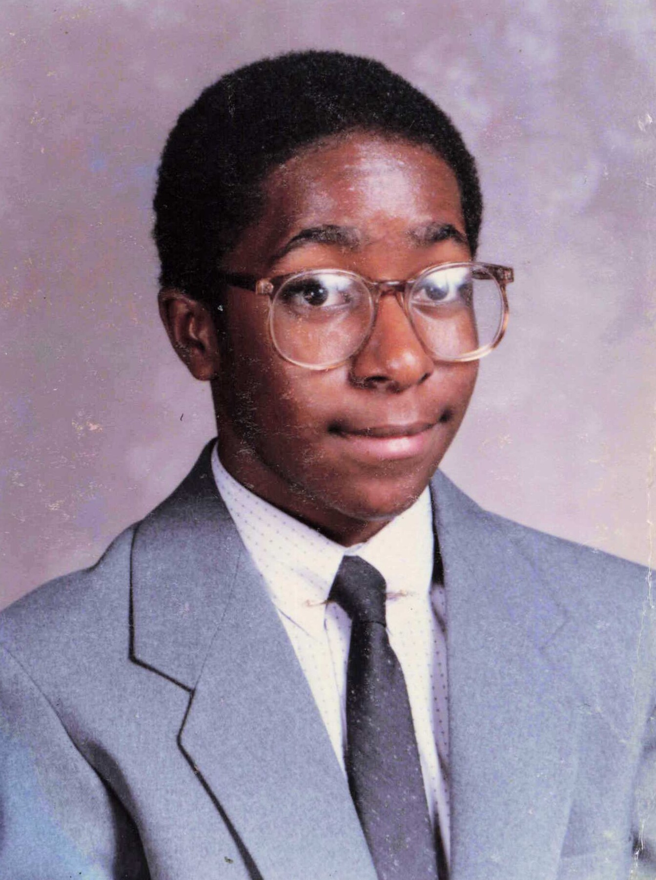 Young man in a light blue suit and dark tie poses for a formal portrait, wearing large round glasses.