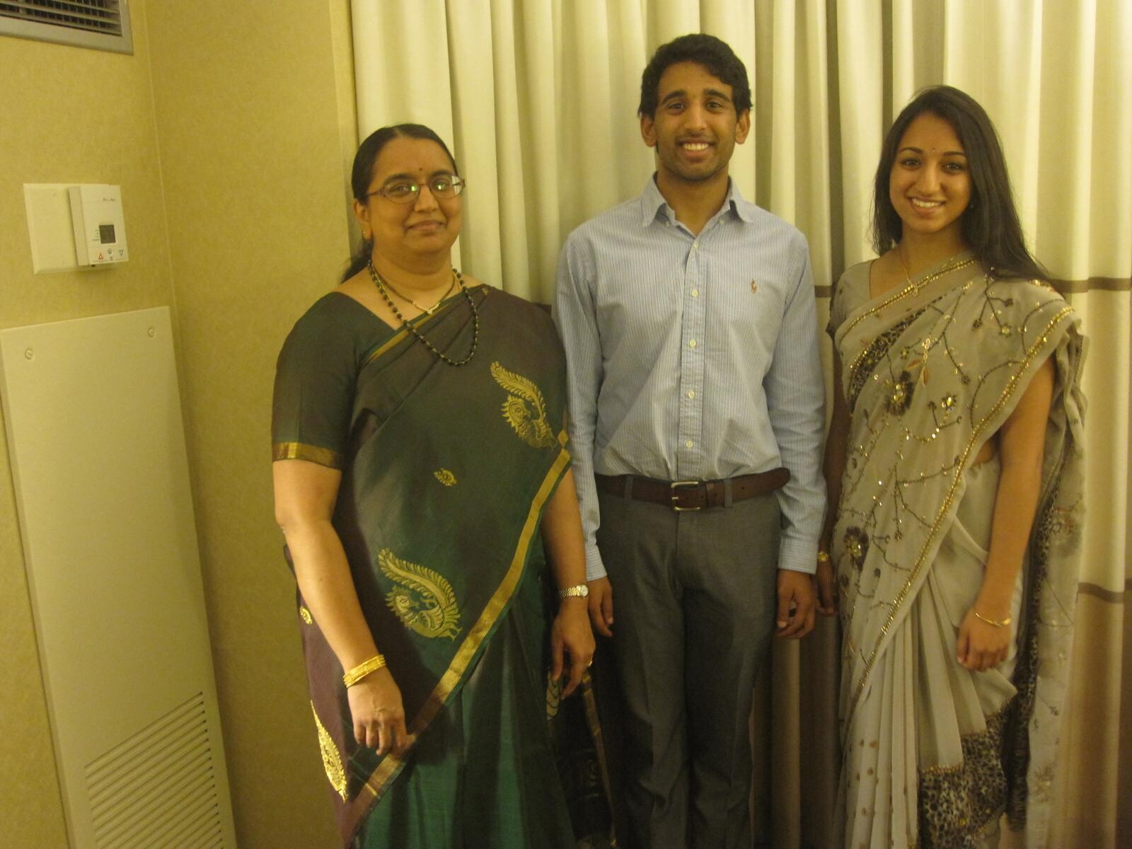 Three people posing indoors in formal attire: woman in green sari, man in light blue shirt, woman in gray beaded sari.