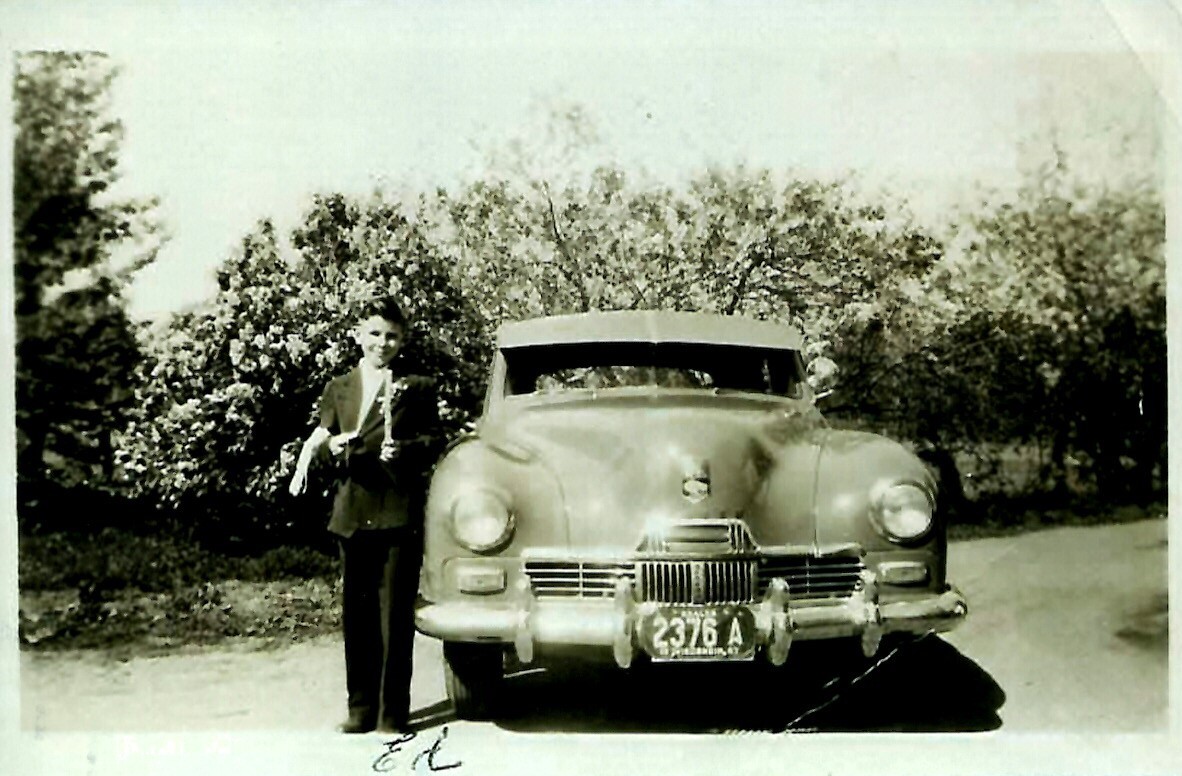 Man in a suit standing beside a vintage mid-century car on a sunny street.