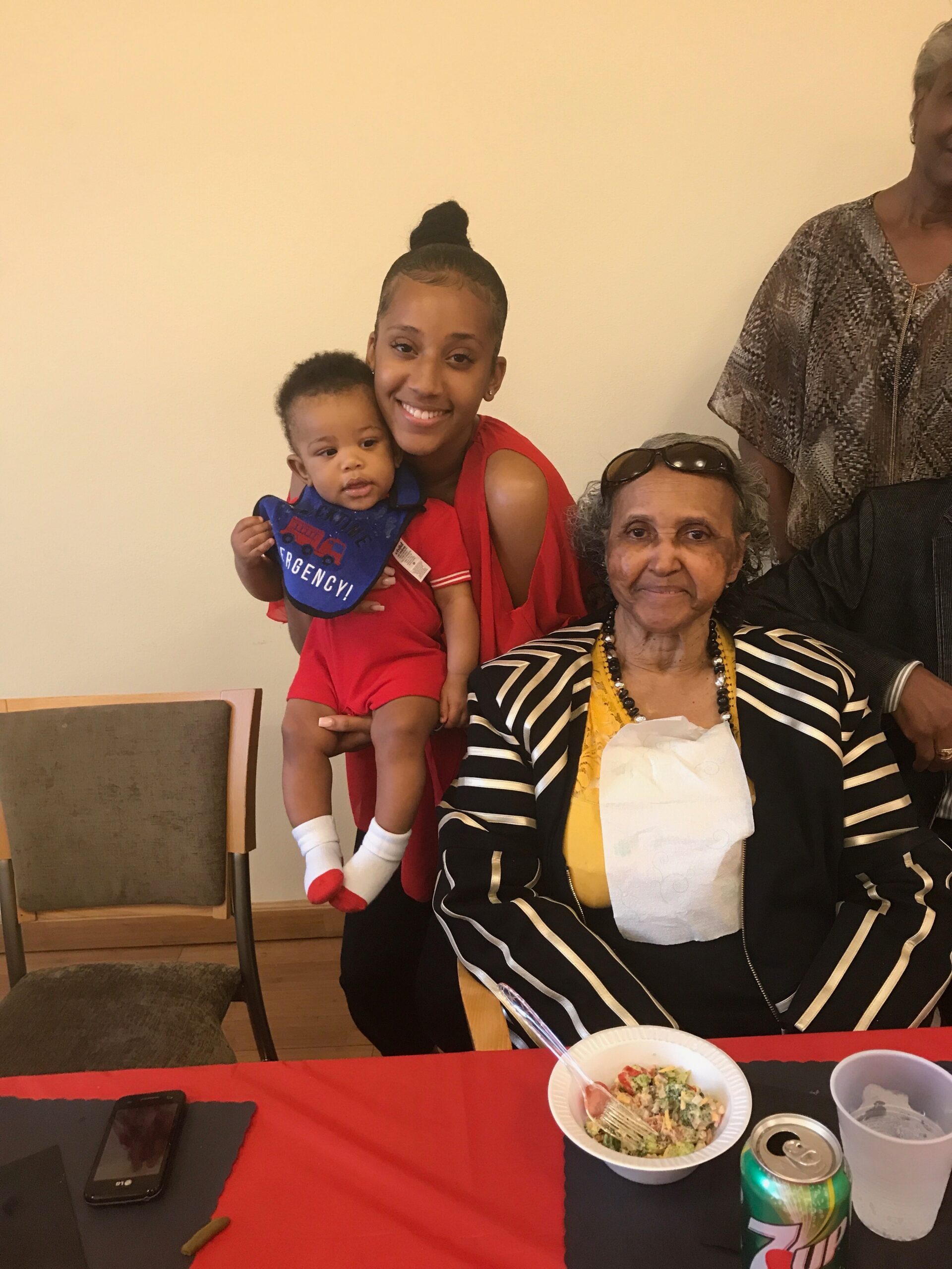Smiling young woman in red holds a baby wearing a blue bib; an elderly woman in striped cardigan sits nearby at a table with food and drinks.