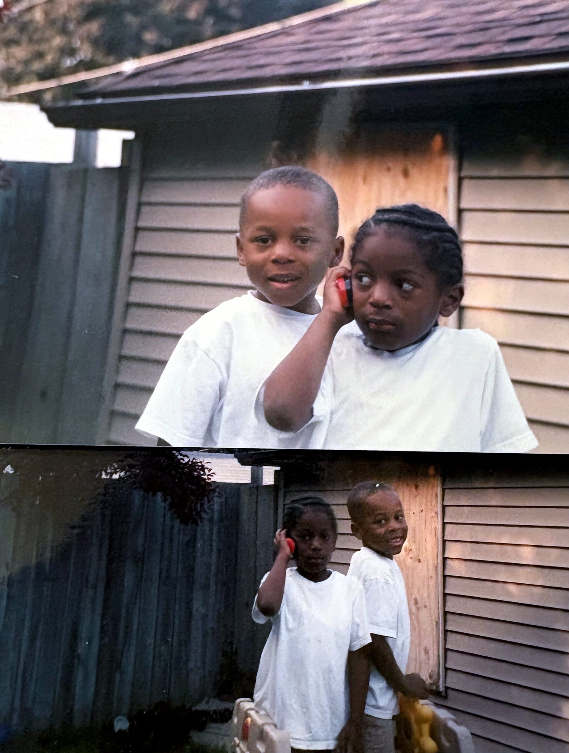 Two children in white shirts stand outside a wooden shed; one smiles at the camera while the other holds a red toy phone nearby.