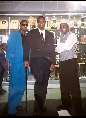 Three men pose in front of a trophy case; left man wears a bright blue suit and sunglasses with a cane, center man in a black suit, right man in a gray vest and hat.