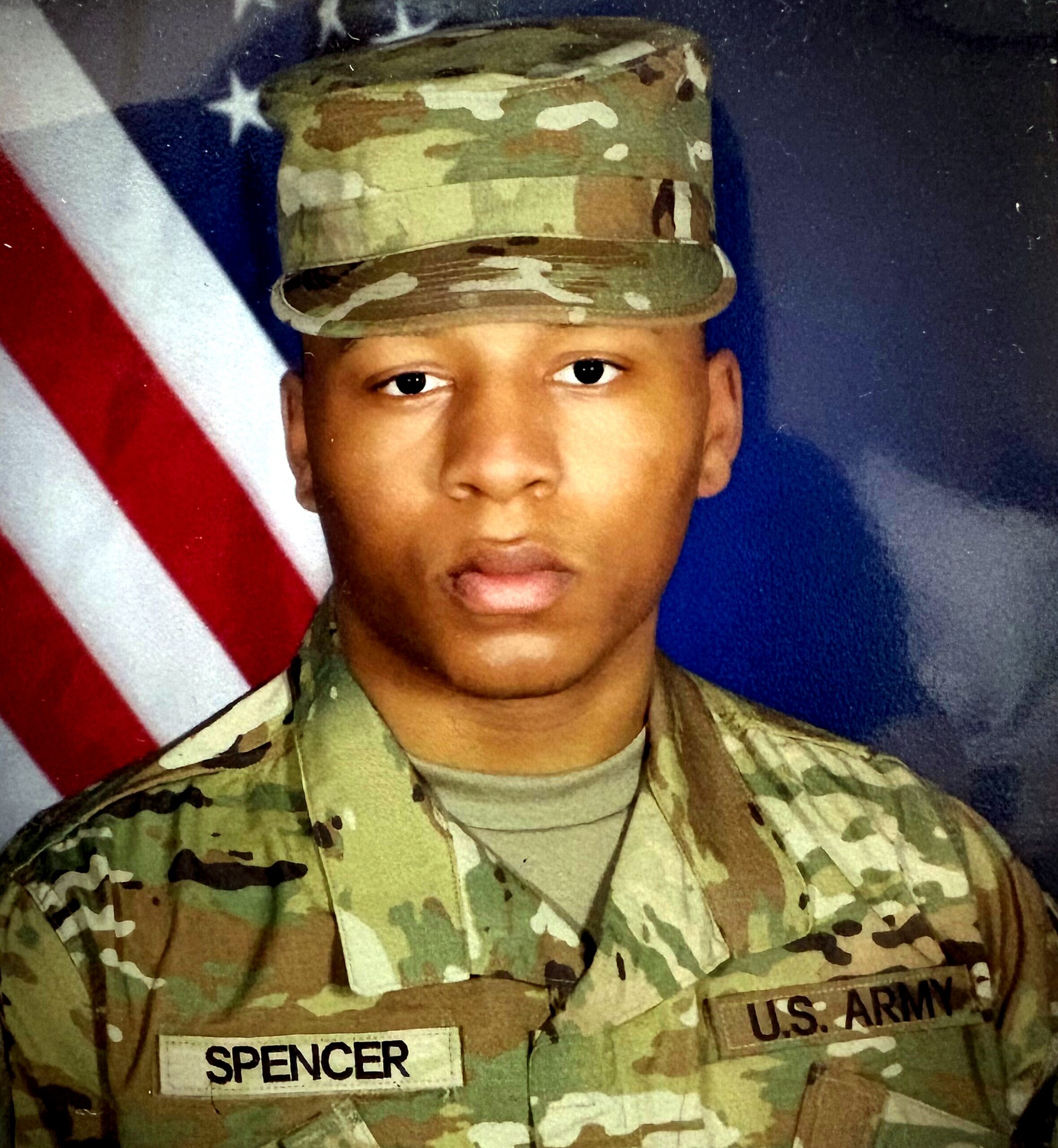 Portrait of a U.S. Army soldier in camouflage uniform, with name tag reading 'SPENCER' and an American flag in the background