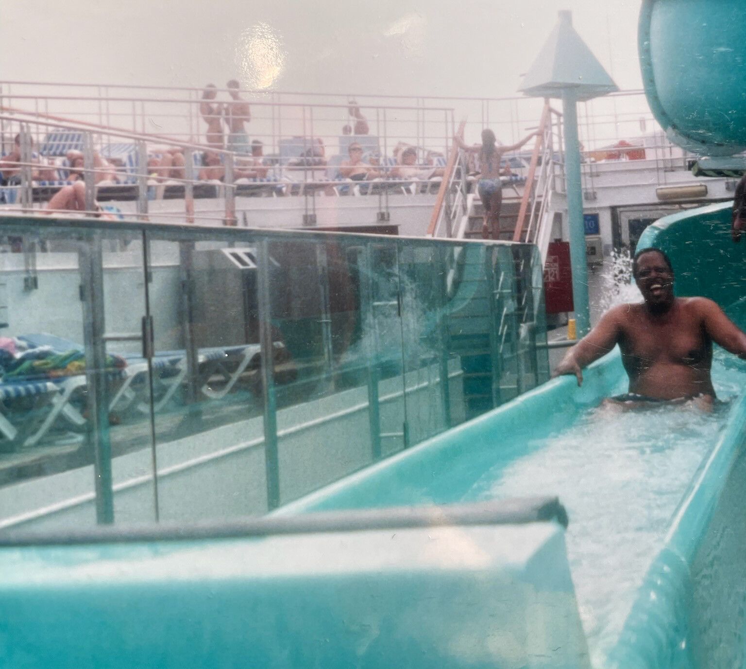 Man shirtless on a turquoise water slide on a cruise ship, smiling as he slides into the pool while deck chairs and people lie in the background.