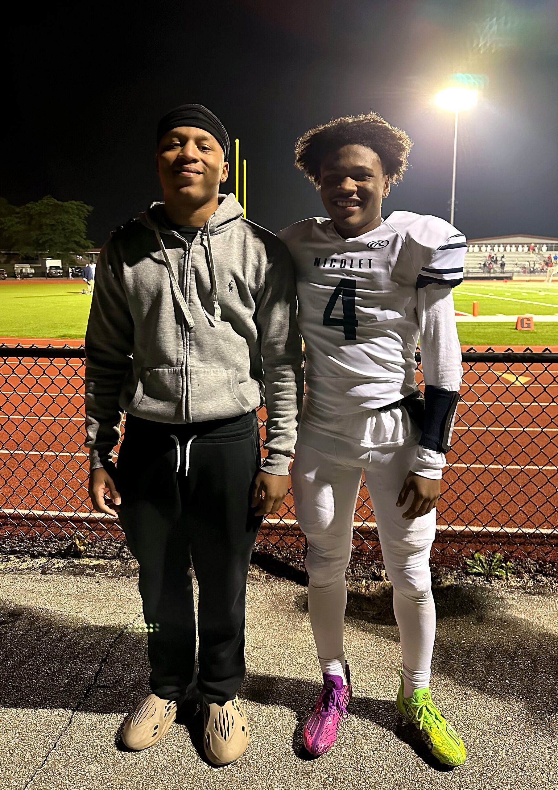 Two young men pose together at a night football field; one in a gray hoodie and tan slides, the other in a white football uniform with number 4 and bright cleats.