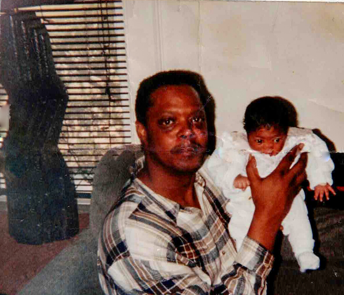 Man in a plaid shirt holding a baby in white clothing indoors near a window with blinds.