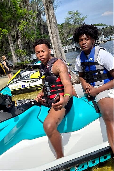 Two young men in life jackets sit on a turquoise jet ski by a dock, posing for the photo as water and trees surround them.