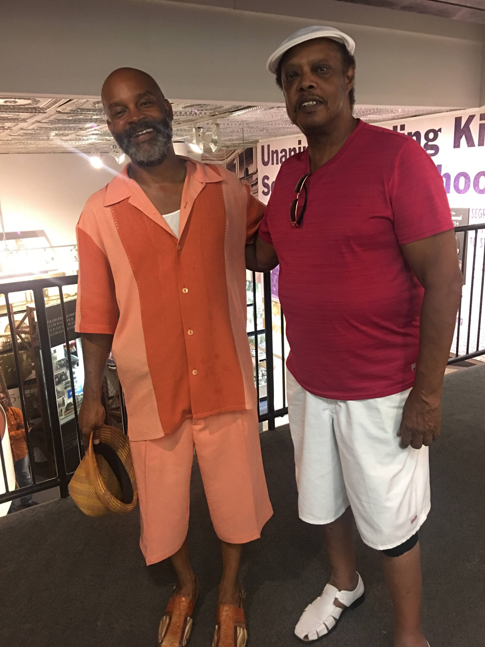 Two men pose together indoors: one in a peach-orange suit and sandals, the other in a red T-shirt and white shorts with sunglasses hanging from his shirt.