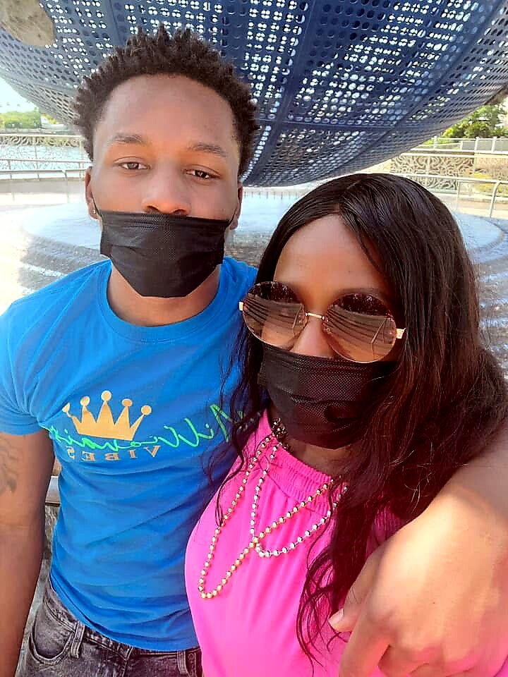 Two people take a close-up selfie wearing black masks, standing in front of a blue sculpture and fountain outdoors.