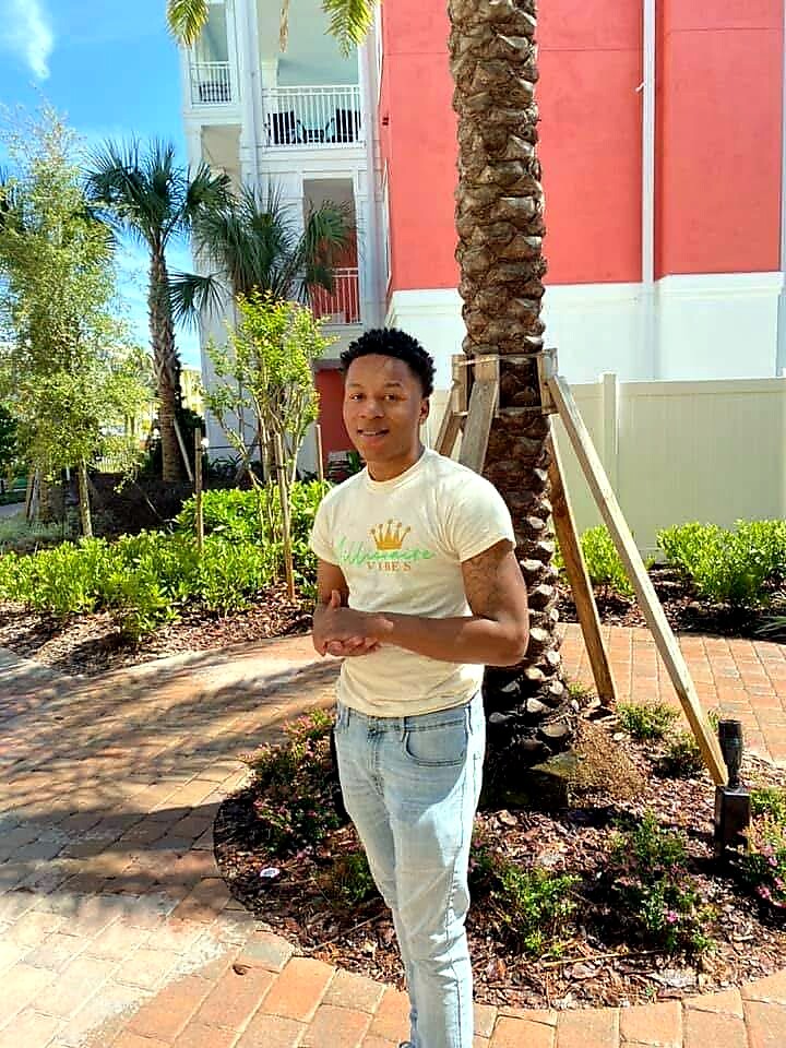 Young man wearing a light-colored T-shirt and jeans stands on a brick walkway in a sunny palm-filled courtyard with residential buildings in the background.