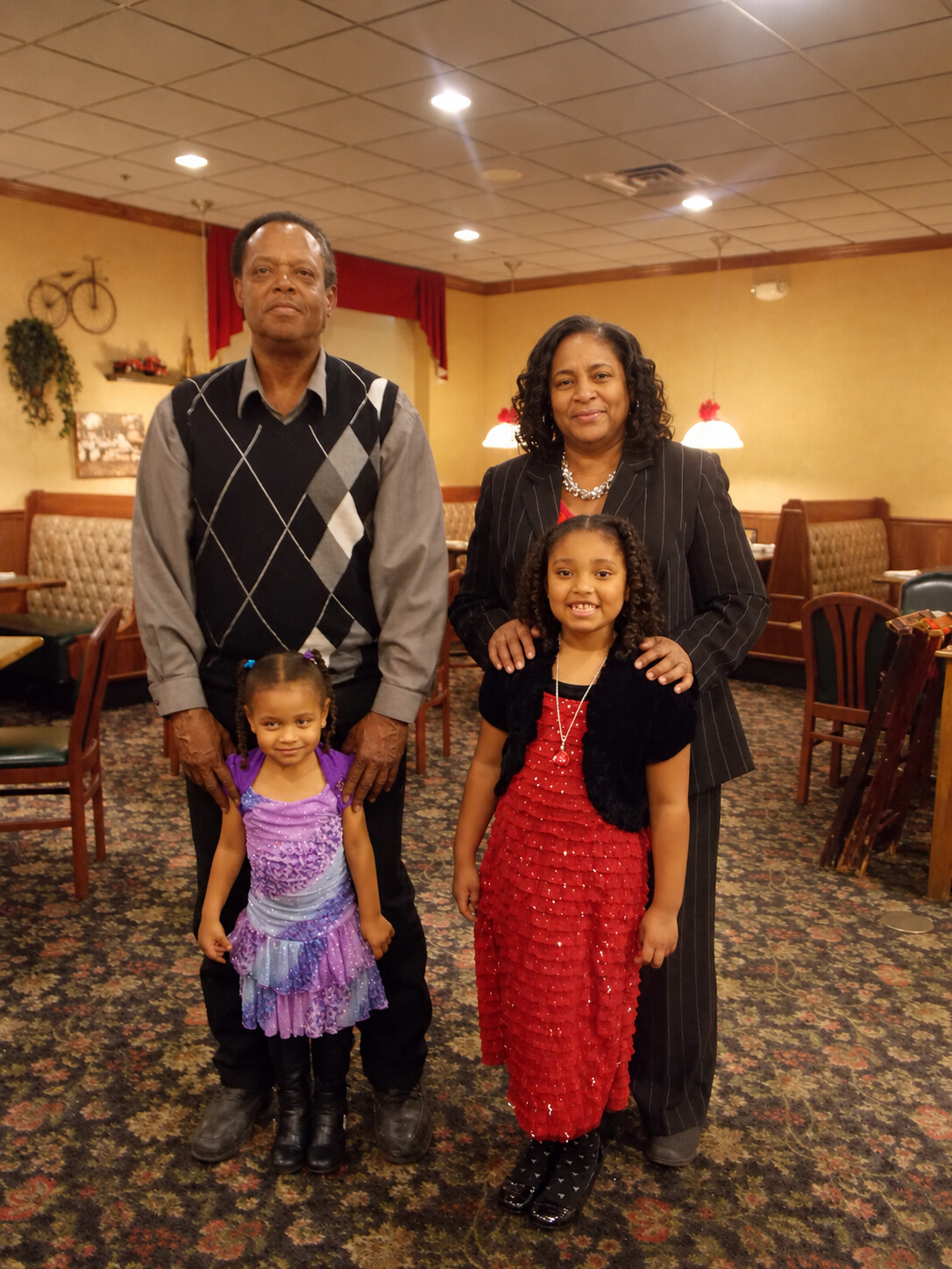 A family of four posing for a photo in a restaurant, two adults with two young girls in colorful dresses.