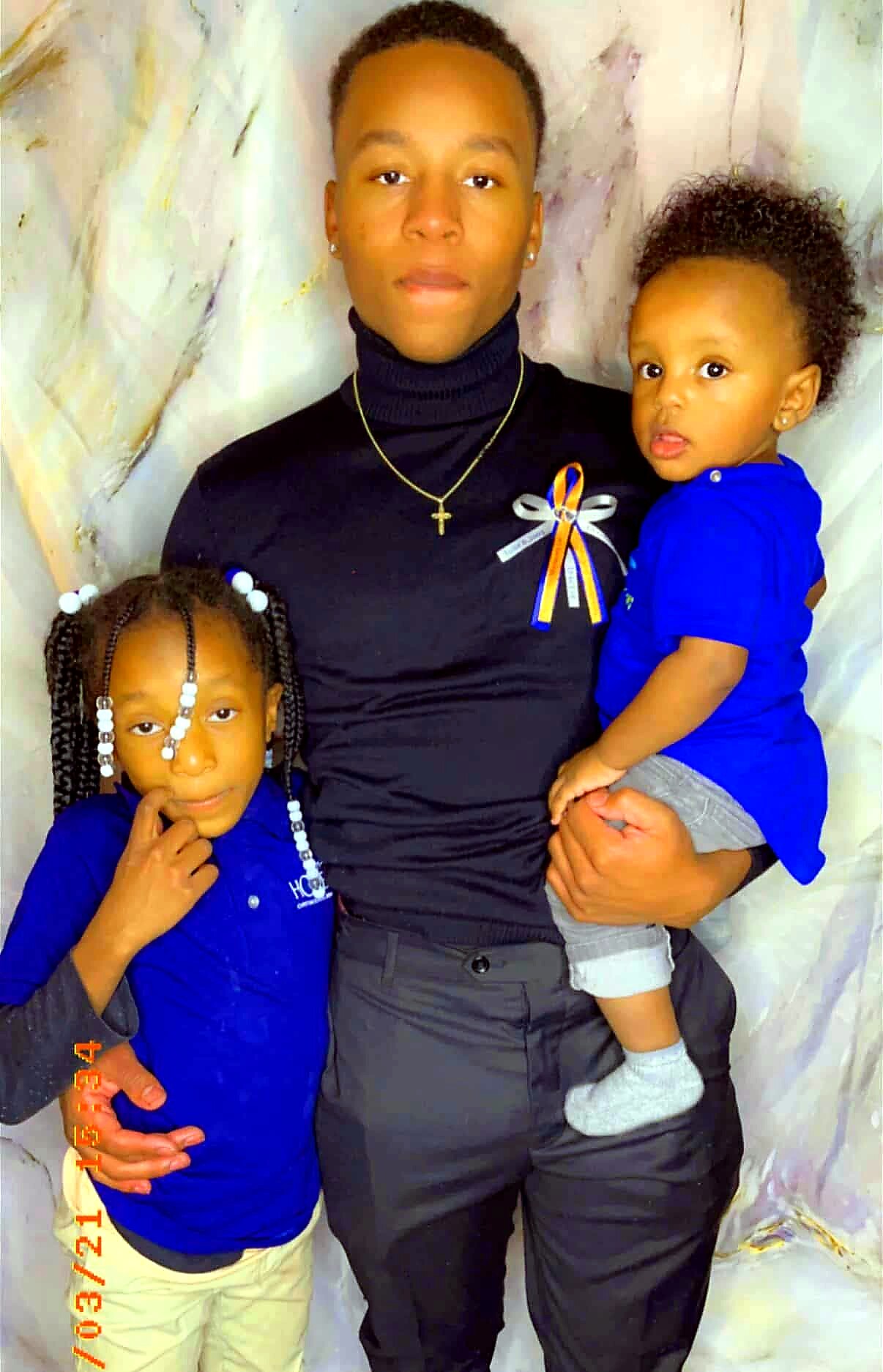 Father in a black turtleneck holds a toddler while his older daughter stands beside him; all wear blue outfits and a ribbon is pinned to his chest.