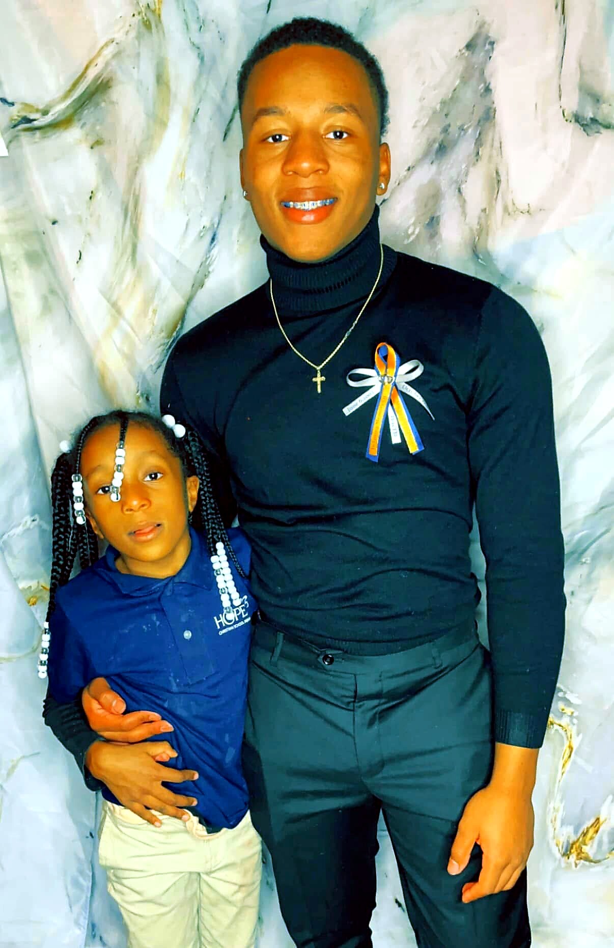 Two people posing together: an adult man in a black turtleneck with a blue-yellow ribbon and a cross necklace, smiling beside a young girl in a blue shirt with beaded braids.