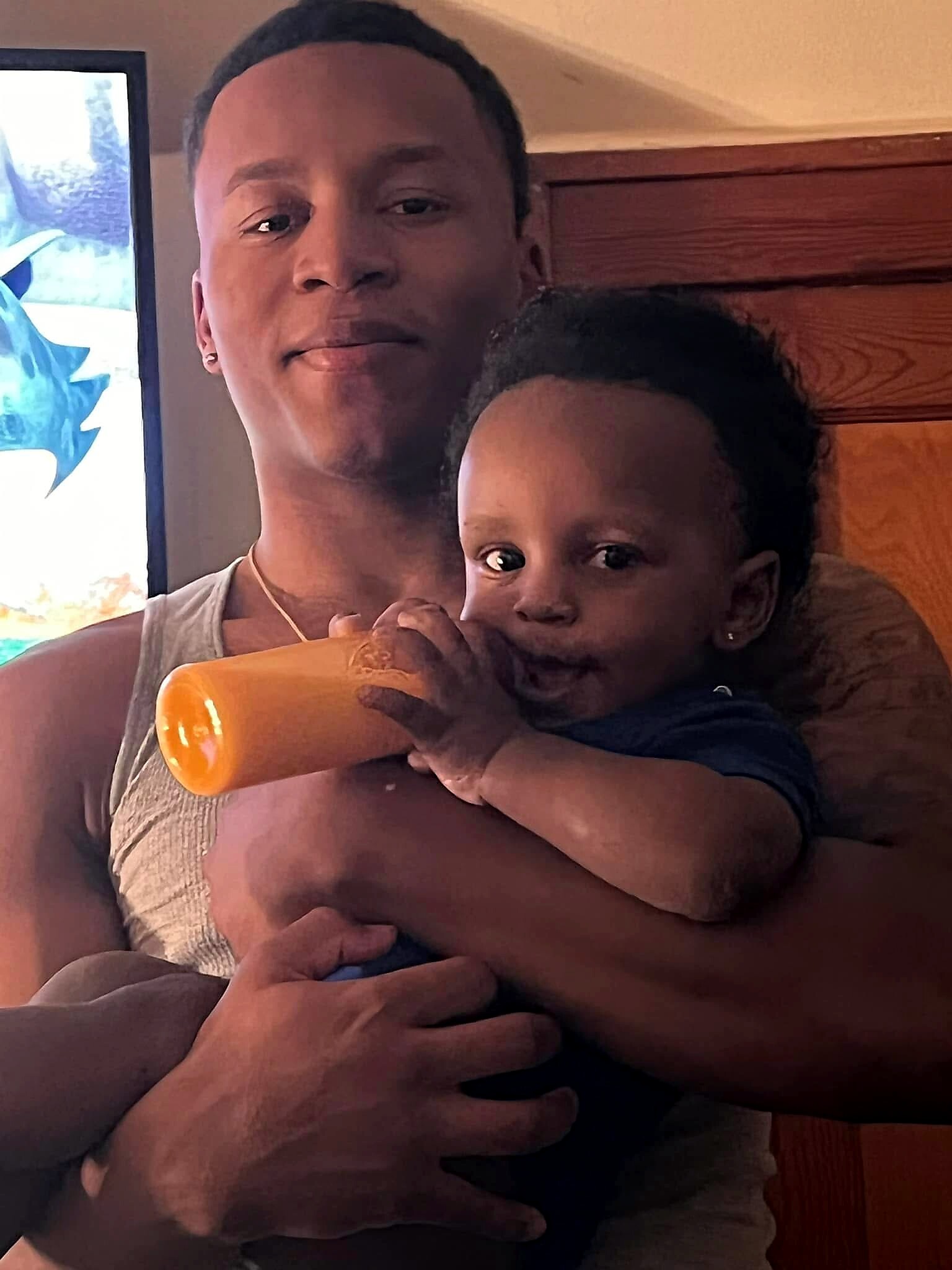 A woman holding a baby who is drinking from an orange bottle, close up from the chest up in a daytime setting.