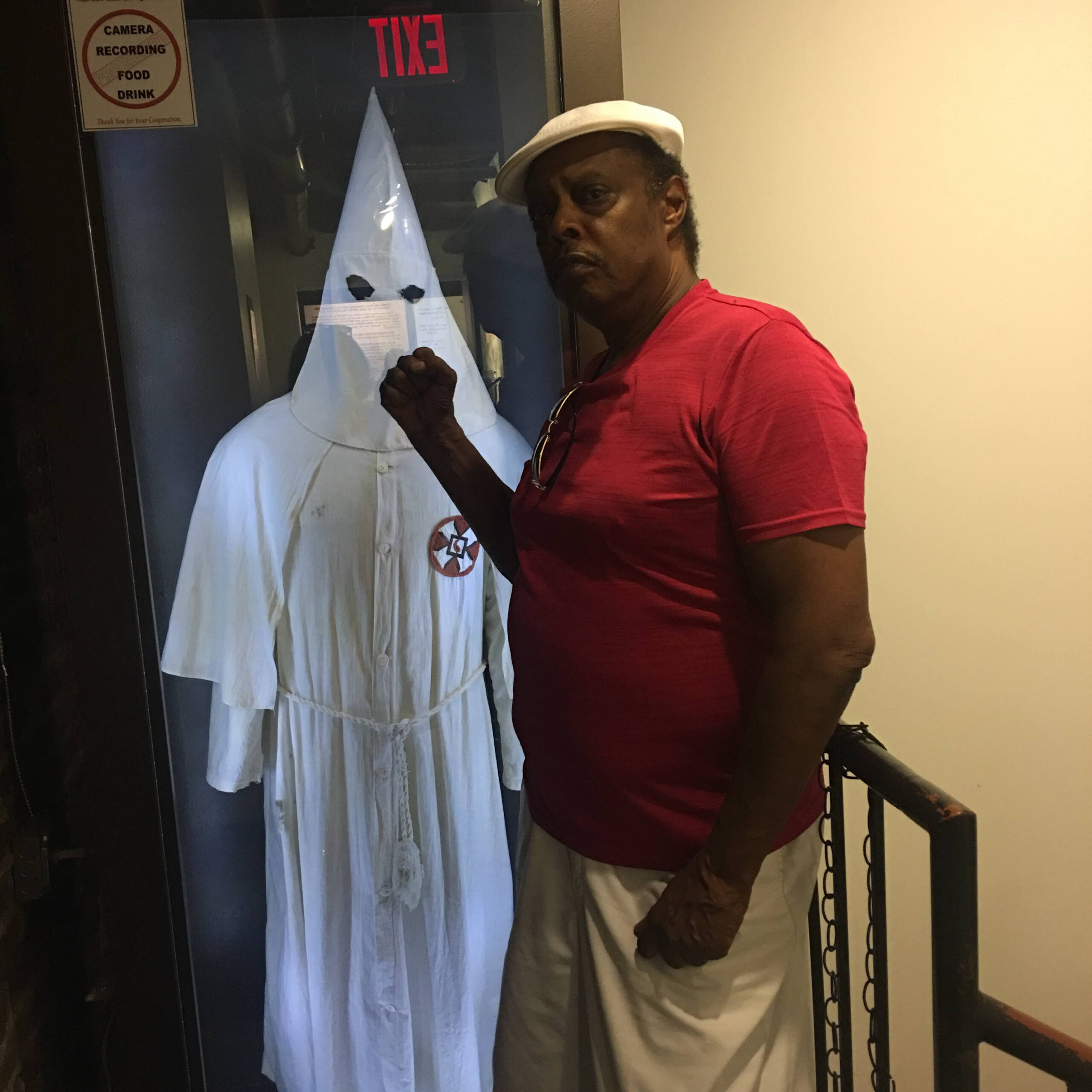 Man in a red shirt and white cap posing next to a person in a white ghost costume behind a glass door; EXIT sign visible above.