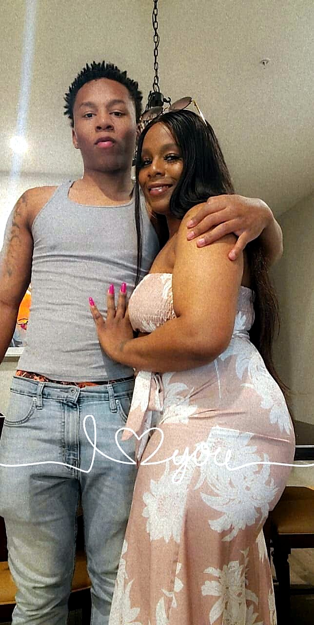 Two friends pose closely indoors; a man in a gray sleeveless shirt and jeans stands beside a woman in a pink floral dress, with the woman resting her hand on his chest and both smiling slightly.