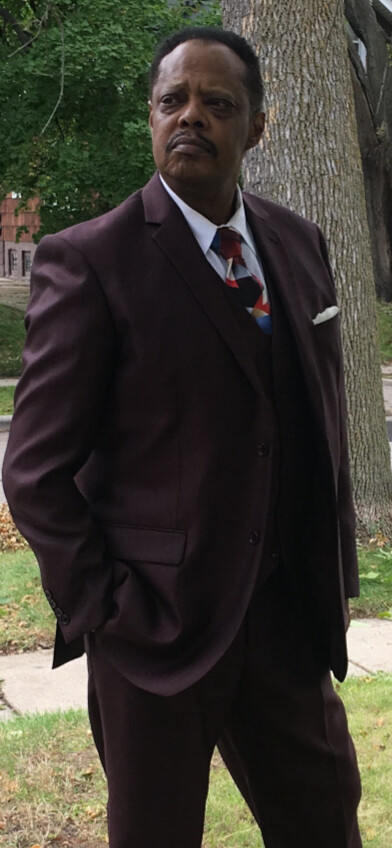 Man in a burgundy suit and vest, with a patterned tie and white pocket square, standing outdoors beside a tree and looking to the side with a serious expression.