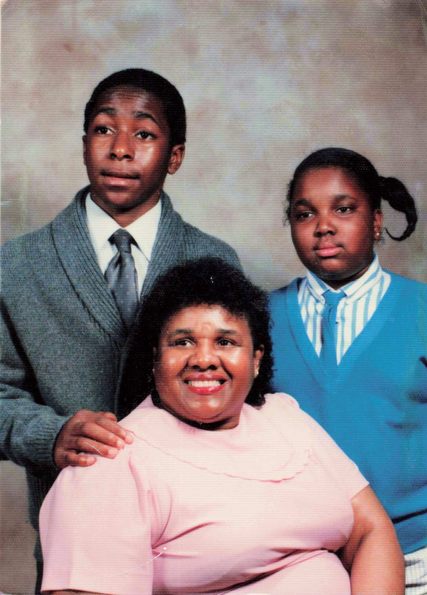 Smiling trio in a studio portrait: a woman in pink in the foreground, a man in a gray suit on the left, and a girl in a blue blazer on the right behind them.