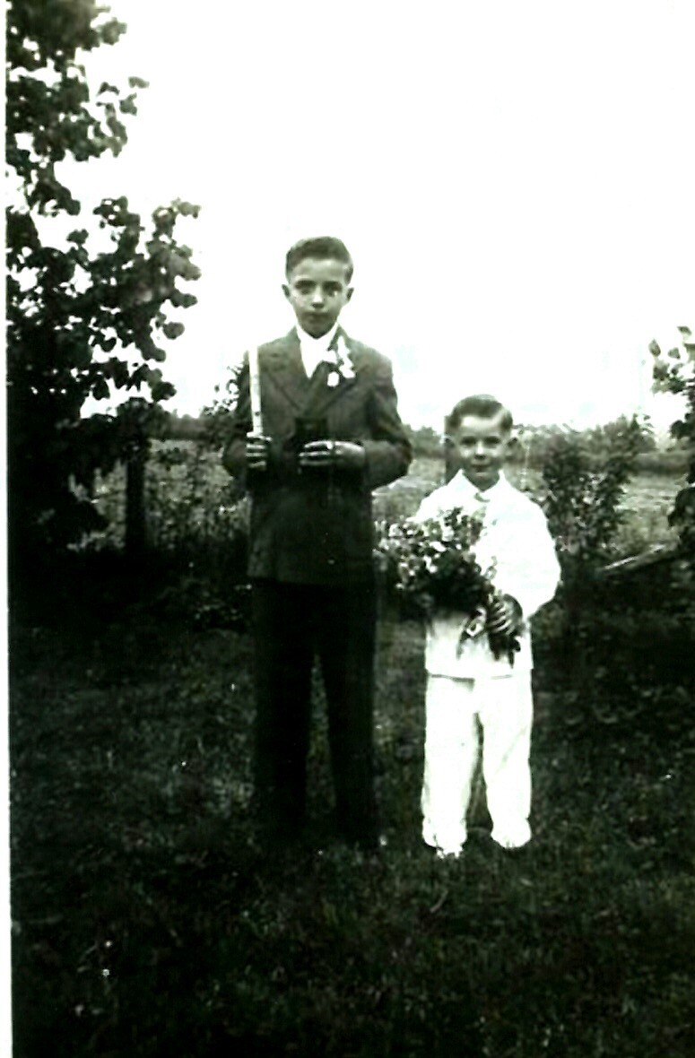 Two boys stand in a garden: older boy in a suit holding a tall candle, younger boy in white holding a bouquet of flowers.