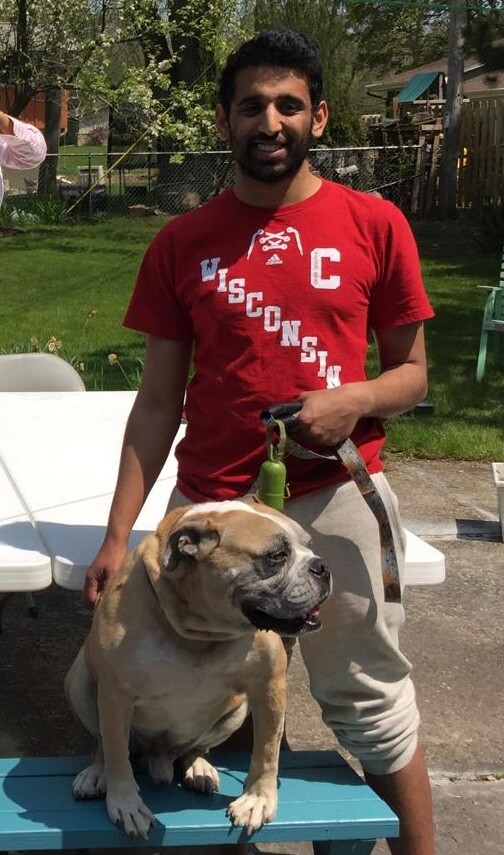 Man in a red Wisconsin shirt stands outdoors, holding a leash attached to a bulldog sitting on a blue bench in a backyard.