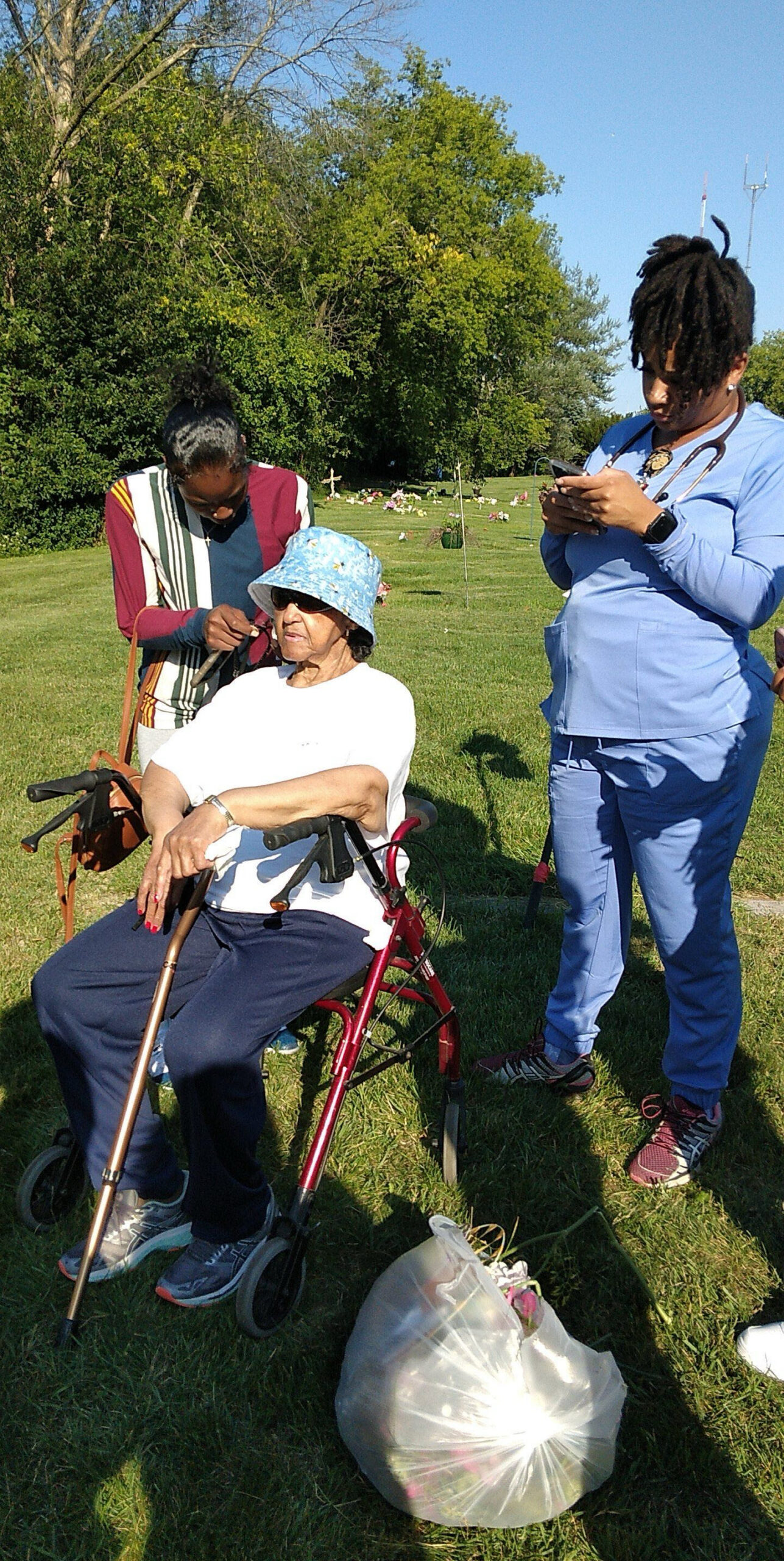 Elderly woman in a white shirt and blue hat sits in a red walker-style wheelchair while a caregiver trims her hair; another caregiver in blue scrubs stands nearby looking at a phone in a grassy cemetery setting.