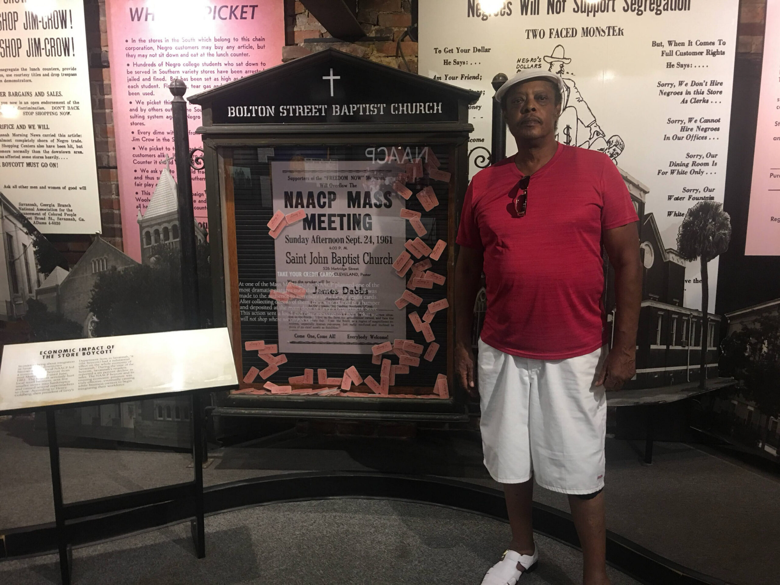 Man in a red shirt and white shorts stands beside a history display featuring a church front and an NAACP Mass Meeting poster with pink sticky notes nearby.