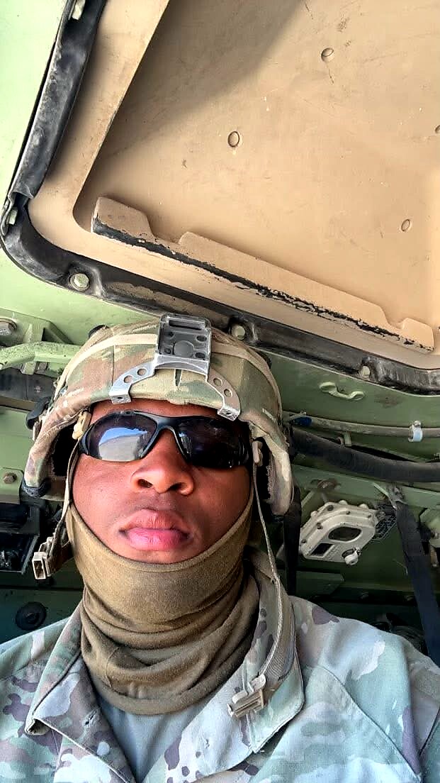 Soldier in camouflage uniform wearing helmet and sunglasses, taking a selfie inside a military aircraft cockpit.