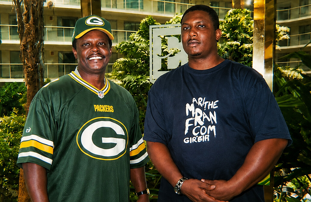 Two men pose together outdoors, one wearing a Packers jersey and cap, the other in a dark graphic T‑shirt.