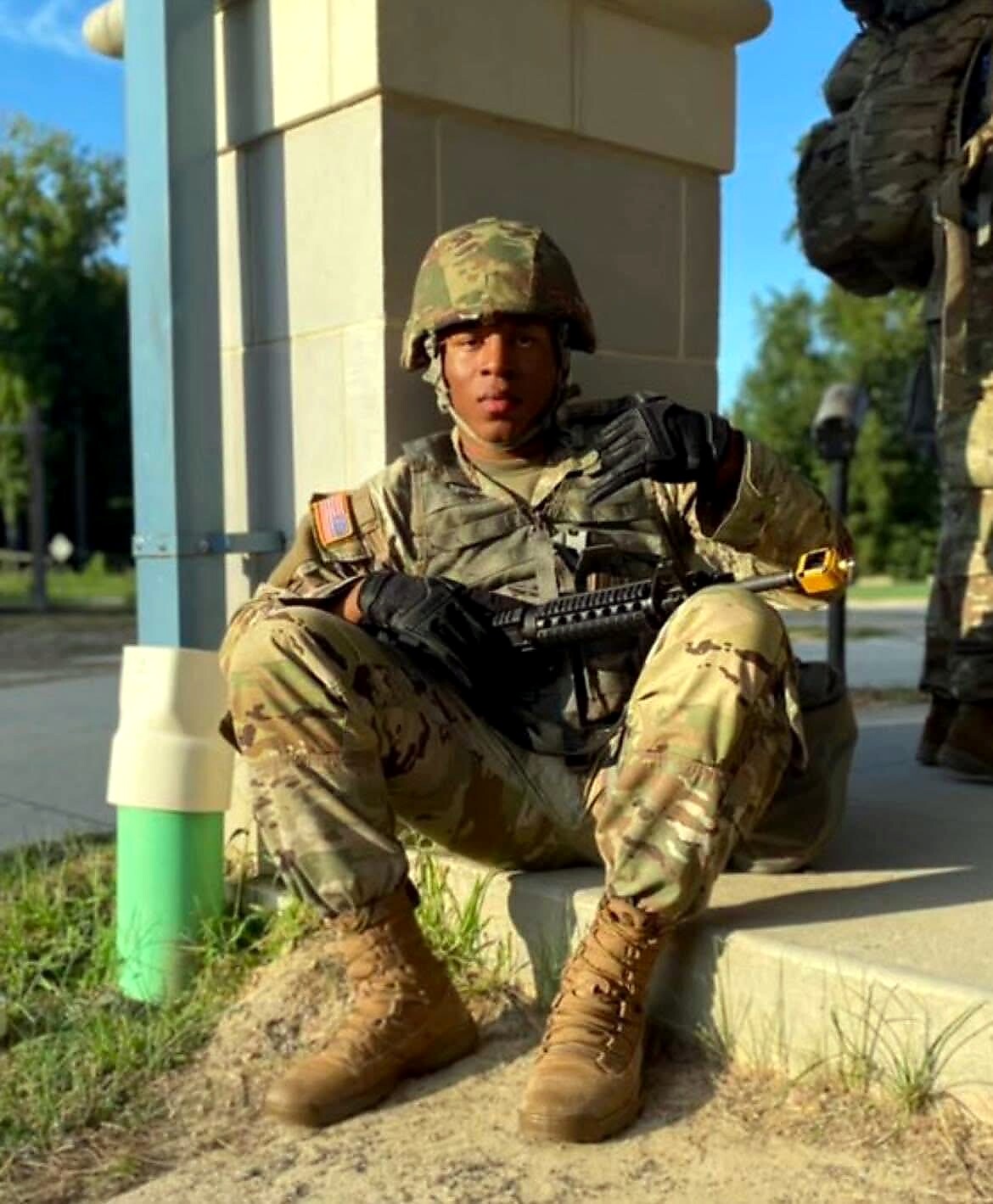 Soldier in camouflage gear and helmet sitting on a concrete step with a rifle across their lap outdoors by a column.