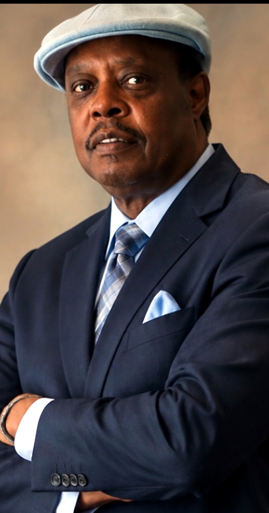 Man in a navy suit, light blue tie and pocket square, wearing a light blue cap, arms crossed in a portrait.