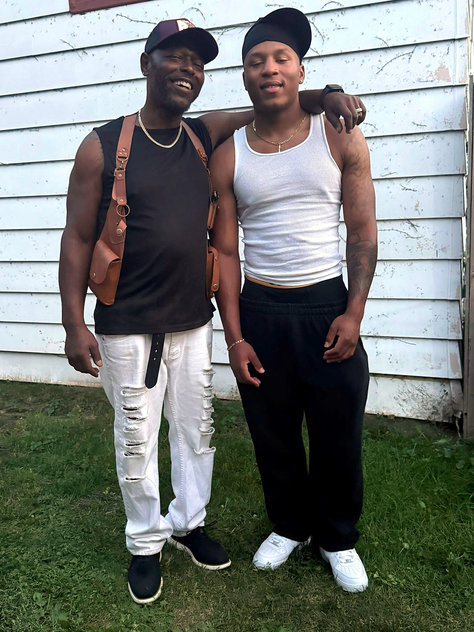 Two men pose arm-in-arm in front of a weathered blue wall; one wears a black sleeveless top with a brown harness, the other a white tank top.