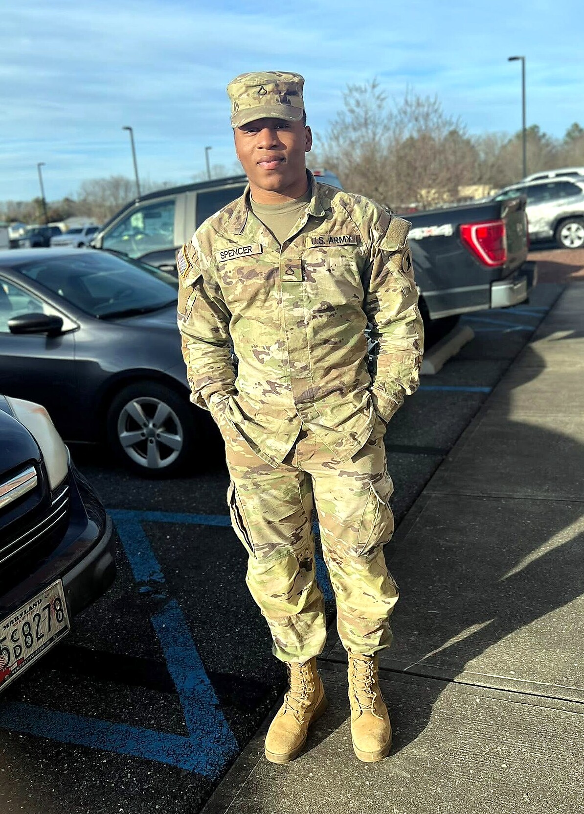 U.S. Army servicemember in camouflage uniform standing with hands in pockets in a parking lot, vehicles in the background.